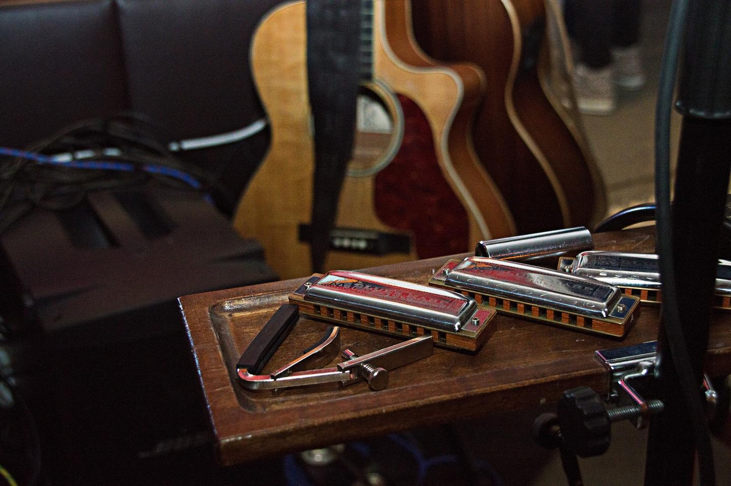 Two diatonic harmonicas resting on a wooden table in a rehearsal room, with acoustic guitars behind them.